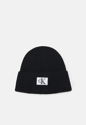 Gorro de punto negro con un dobladillo, que presenta una etiqueta rectangular blanca con un logo negro. Tejido texturizado, forma cilíndrica.