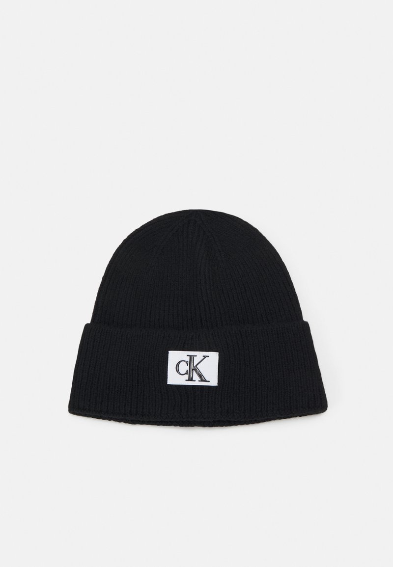 Gorro de punto negro con un dobladillo, que presenta una etiqueta rectangular blanca con un logo negro. Tejido texturizado, forma cilíndrica.