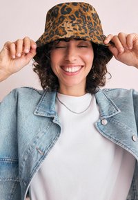 Chapeau seau en imprimé léopard, marron avec une texture lisse. Porté par une personne portant une veste en denim clair sur une chemise blanche.