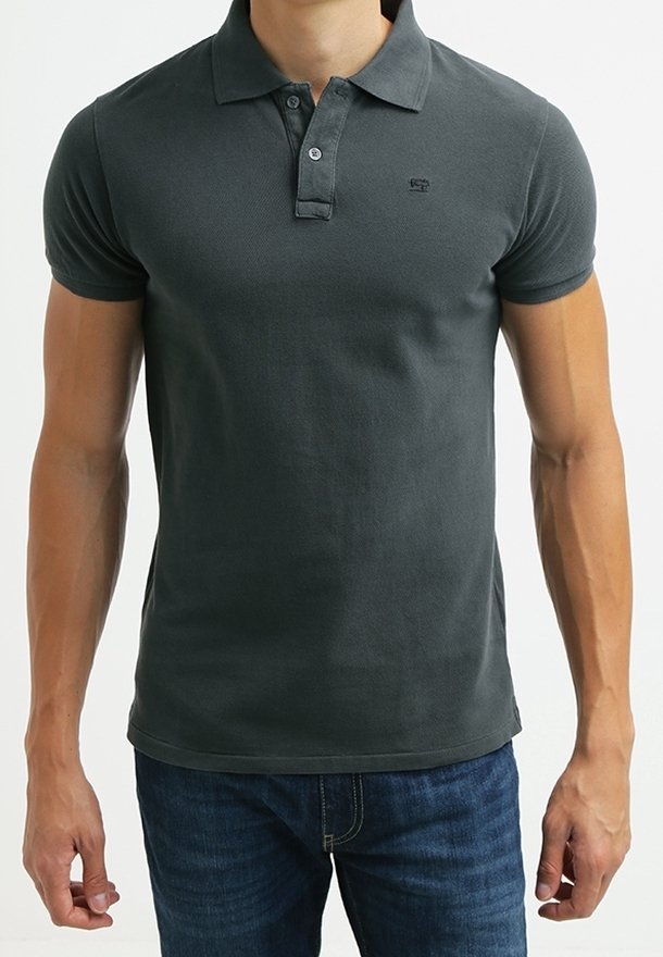 Homme portant un polo gris foncé ajusté avec un col et deux boutons, associé à un jean bleu, debout devant un fond uni.