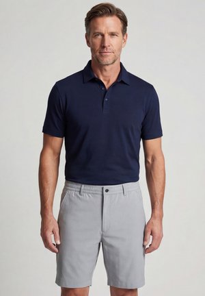 Man standing wearing a navy short-sleeve polo shirt and light gray tailored shorts against plain gray background.