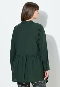 Camicia verde a maniche lunghe con vita arricciata, dotata di colletto e polsini con bottoni. Il tessuto appare morbido e liscio con un design fluido.