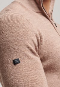 Jasnoróżowy sweter z dzianiny z zamkiem przy kołnierzu, z prążkowanym wykończeniem i małą czarną metką na lewym rękawie.