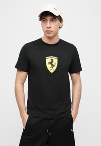 Νέος άνδρας φοράει μαύρο μπλουζάκι Ferrari και μαύρα σορτς Puma, με τα χέρια στις τσέπες και λευκό καπέλο Puma, κοιτάζοντας προς τα πλάγια.