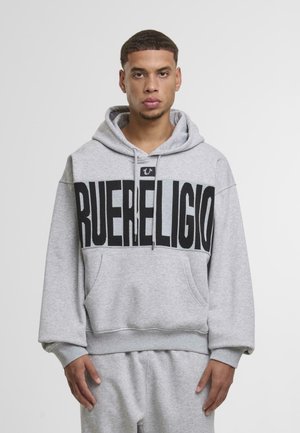 Young man wearing a gray hoodie and matching sweatpants with bold black "TRUE RELIGION" text across the chest.