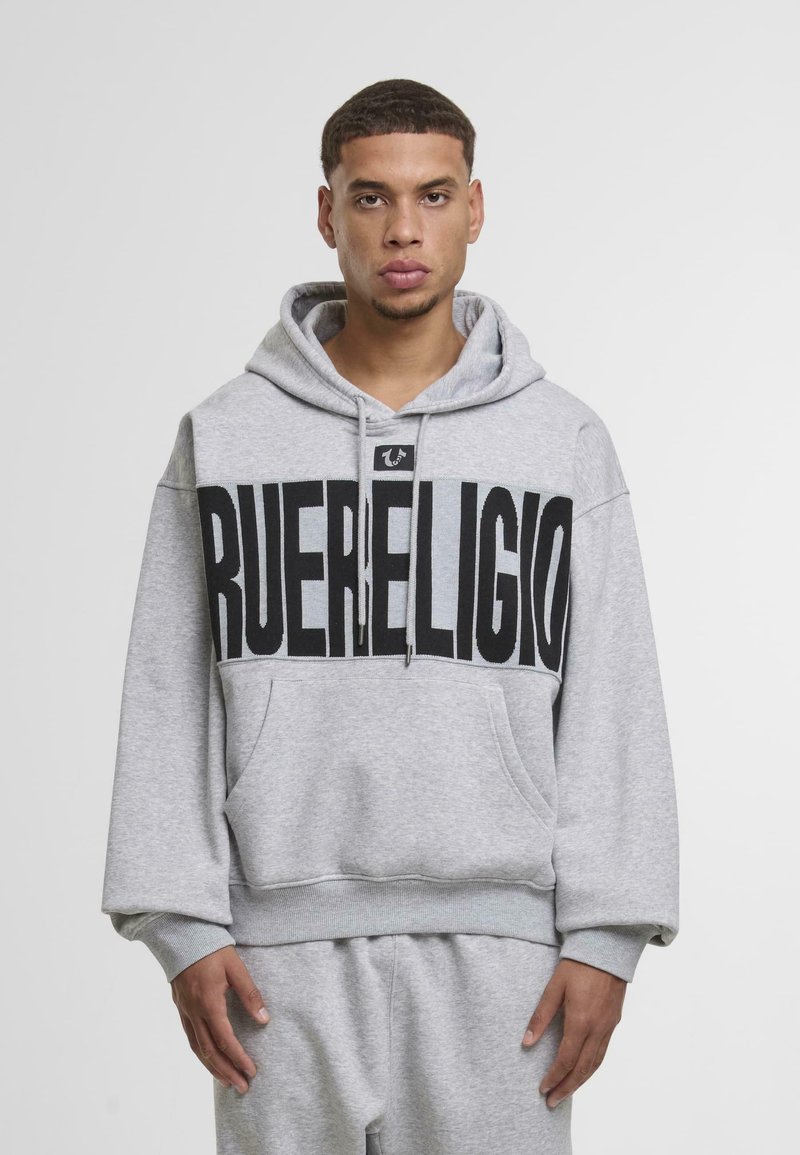 Giovane uomo che indossa un felpa grigia con cappuccio e pantaloni della tuta coordinati, con il grande testo nero "TRUE RELIGION" sul petto.