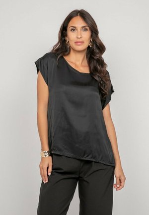 Femme aux cheveux longs et foncés portant un chemisier satiné noir à manches courtes et un pantalon noir, debout devant un fond clair uni.