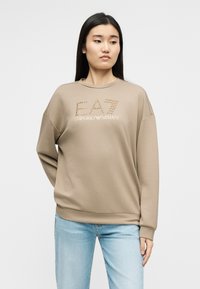 Νέα γυναίκα με μακριά σκούρα μαλλιά φοράει μπεζ φούτερ Emporio Armani και ανοιχτόχρωμο τζιν, κοιτάζοντας ελαφρώς προς τα αριστερά της.