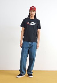 Fekete pamut póló fehér és piros Reebok logóval, laza kék farmernadrággal és fekete slip-on cipővel párosítva.