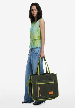 Green canvas tote bag with contrasting light green straps and a front zipper pocket. Features a brown logo patch and a spacious design.