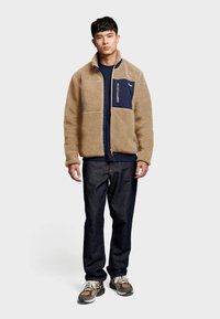 Tan fleece jacket with navy pockets, zip closure, and soft texture. Worn over a dark shirt and paired with dark jeans and sneakers.