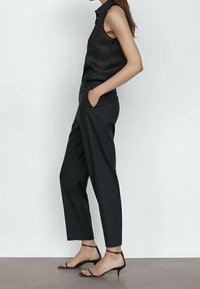 Blouse noire sans manches avec un col montant, associée à un pantalon noir fuselé. Le modèle porte des talons noirs à lanières. Tissu lisse, design minimaliste.