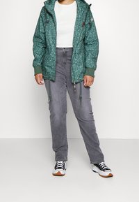 Green patterned jacket with adjustable hood, paired with gray pants and black sneakers featuring white accents and brown soles.