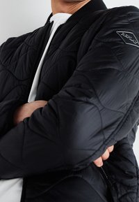 Giacca bomber nera trapuntata con zip frontale, polsini a coste e una toppa con logo sulla manica. Il tessuto ha una superficie liscia.
