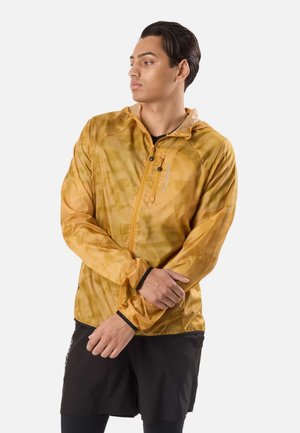 Jonge man die de mouw van een mosterdgele jas met ritssluiting en capuchon omhoog schuift, draagt zwarte korte broek en legging, kijkt naar links.