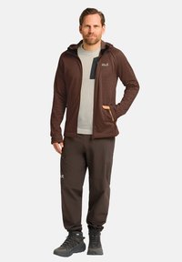 Bruine zip-up jas met een capuchon, gecombineerd met een lichtgrijs shirt en bruine pantalons. De jas heeft zakken en een textuurstof.