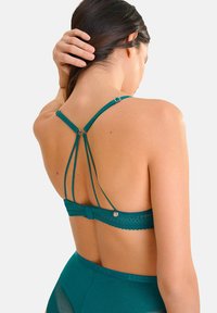 Bralette en dentelle teal avec de fines bretelles réglables, une bande texturée et un détail décoratif en matériel circulaire à l'arrière.