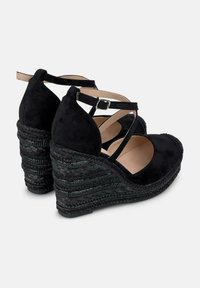 Scarpe con zeppa in suede nero con design a strappo incrociato, tacco intrecciato testurizzato e punta chiusa. Presentano una cinghia alla caviglia regolabile e una soletta imbottita.