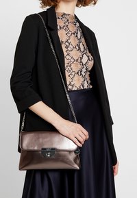 Metalisk bronze håndtaske med en glat overflade, rektangulær form og lås. Har en kædetræk og holdes af en person.