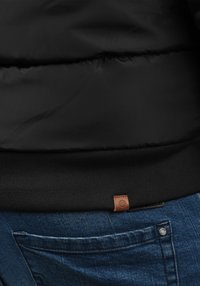 Zwarte donzen jas met een geribbelde zwarte zoom en een bruin leren logolabel. Draagt blauwe spijkerbroek met een zakdetail.