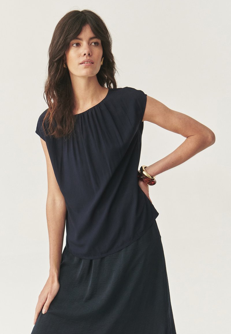 Blusa blu navy con maniche a cappuccio, caratterizzata da un collo arricciato e una silhouette fluida, abbinata a una gonna di tessuto scuro.