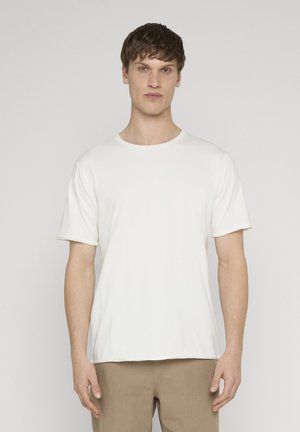 Giovane uomo con capelli castani corti che indossa una t-shirt bianca semplice a maniche corte e pantaloni beige, in piedi davanti a uno sfondo grigio chiaro.