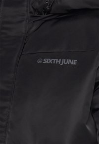 Giubbotto a vento nero con una texture liscia. Presenta una tasca con zip e il logo stampato sul petto. Materiale leggero e isolato.
