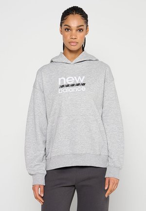 Donna con capelli intrecciati che indossa una felpa con cappuccio New Balance grigio chiaro e pantaloni grigio scuro, in piedi davanti a uno sfondo semplice.