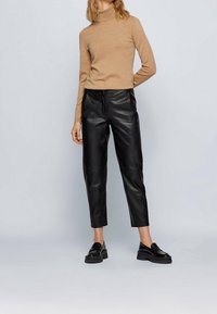 Top de cuello alto beige combinado con pantalones de cuero negros de cintura alta. Los pantalones tienen un corte ajustado, textura suave y llegan justo por encima del tobillo. Mocasines negros completan el conjunto.