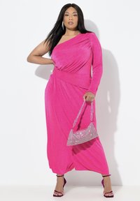 Różowa sukienka na jedno ramię z drapowanym materiałem i długim rękawem. Do niej dobrana błyszcząca torebka oraz sandały na obcasie z paskami i odkrytymi palcami.