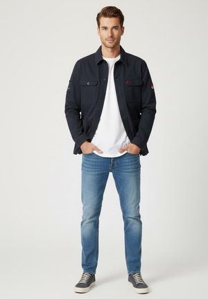 Chaqueta azul marino oscura con botón delantero y bolsillos, combinada con una camiseta blanca y jeans de color azul claro, completada con zapatillas grises.