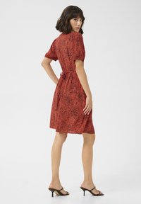 Robe rouge avec un motif tacheté noir, manches courtes bouffantes, taille cintrée avec un lien, coupe au genou et texture de tissu lisse.