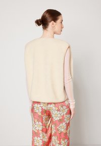 Crème-kleurige fleece vest met een losse, mouwloze pasvorm. Het model draagt een broek met een patroon in roze met grote bloemenmotieven.