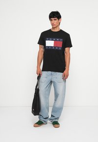 Mustmust värvi T-särk, millel on ristkülikukujuline logo, kus on kirjas "TOMMY JEANS" valge ja punasega tumeläänil, koos helesiniste teksade ja roheliste sneakeritega.
