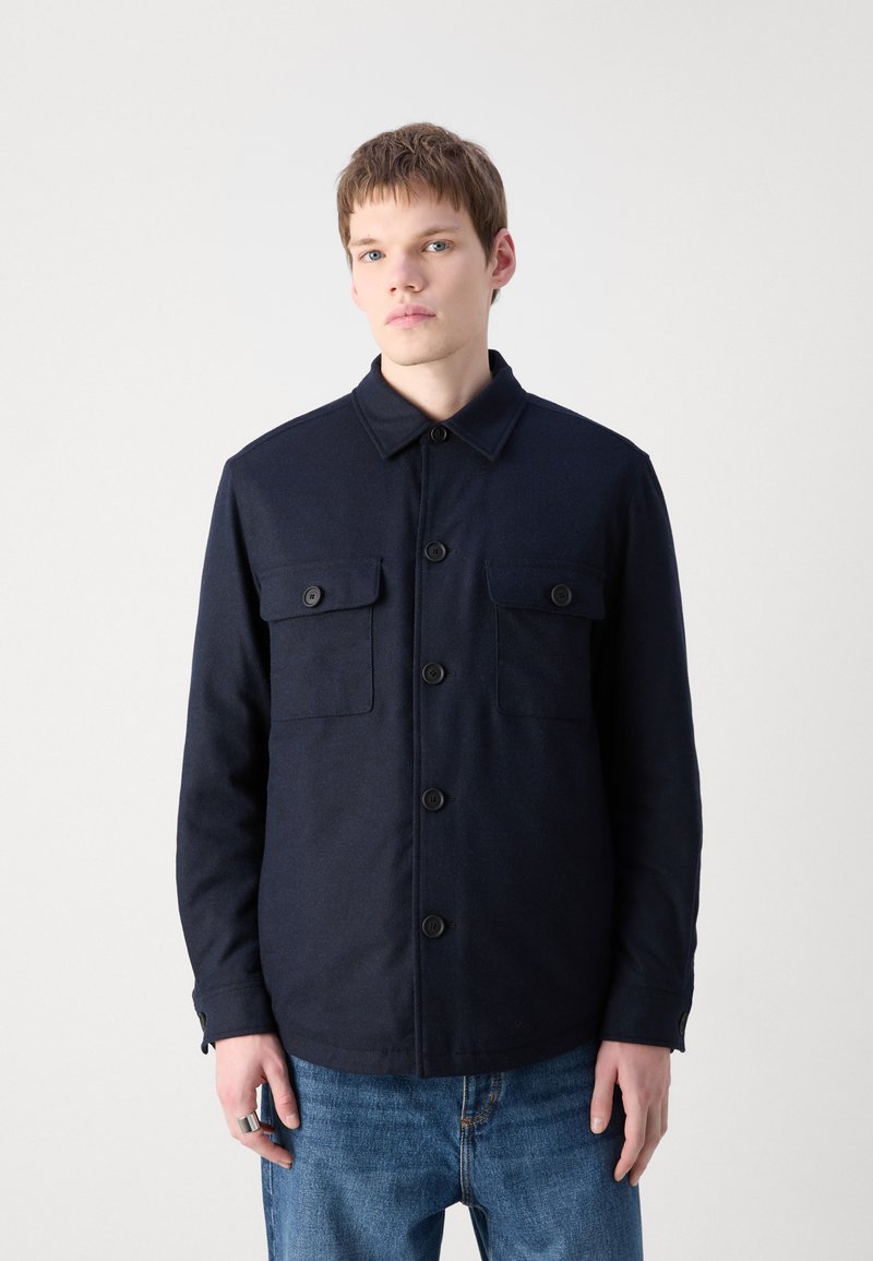 Veste bleu marine à boutons, avec col, deux poches poitrine et manches longues. Fabriquée dans un tissu texturé avec une coupe droite.