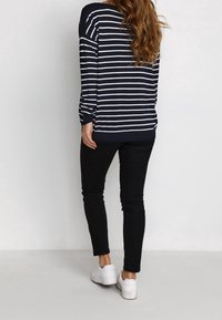 Gestreifter Langarmpullover in Marineblau und Weiß, lockere Passform, kombiniert mit schwarzen, eng anliegenden Hosen und weißen Sneakers, von hinten betrachtet.