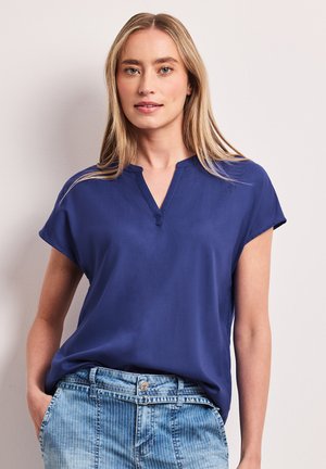 Blondynka stoi na neutralnym tle, ubrana w niebieską bluzkę z krótkim rękawem i jasnoniebieskie dżinsy w cienkie paski, z rękami w kieszeniach.