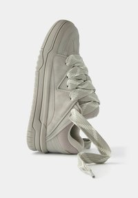 Hellgrauer Sneaker mit dicker Sohle und extra breiten, locker gebundenen Schnürsenkeln, der auf einer weißen Oberfläche liegt.