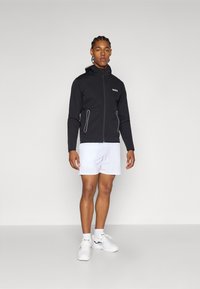 Svart zip-up jacka med vita accenter, långa ärmar och huva; matchad med vita shorts och vita sportskor. Logotyp på bröstet.