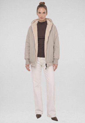 Vrouw draagt een beige capuchonjack met bontvoering, donkerbruine top, witte broek en bruine puntige schoenen, staand tegen een effen achtergrond.