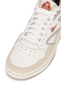 Biele kožené tenisky s šedými akcentmi, suédové béžové prehozy, perforovaná špička, ploché šnúrky a červené logo na jazyku.