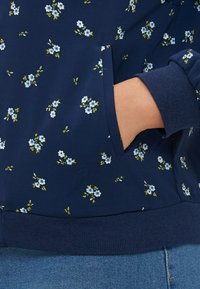 Sweatshirt bleu marine à imprimé floral avec de petites fleurs blanches et bleues, poignets et ourlet côtelés. Il comporte des poches latérales et une texture douce.