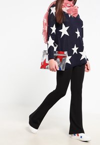 Frau, die einen navyfarbenen Pullover mit weißen Sternen, schwarze Hosen, weiße Turnschuhe mit roten und blauen Sternen, einen pinken Schal trägt und eine silberne Clutch mit Sternenmuster hält.