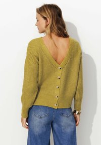 Gilet en tricot vert avec un col en V et des détails de bouton dans le dos. Les manches sont longues et le tissu a une apparence texturée.