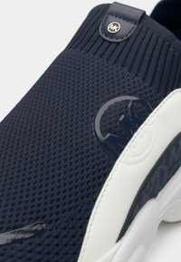 Ναυτικά πλεκτά αθλητικά παπούτσια με υφή στο επάνω μέρος, διαθέτουν λευκή σόλα από καουτσούκ και γυαλιστερή λεπτομέρεια λογότυπου στο πλάι, με ριμπ περιλαίμιο.