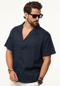 Kurzärmliges, marineblaues Hemd mit einem strukturierten, floralen Muster, Knopfverschluss und einem entspannten Kragen. Trägt dunkle Sonnenbrillen.