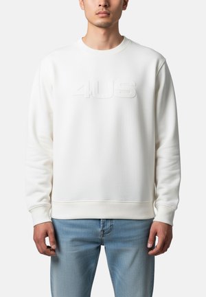 Uomo che indossa una felpa bianca a maniche lunghe con la scritta "4US" in rilievo sul petto e jeans azzurri, in piedi davanti a uno sfondo semplice.