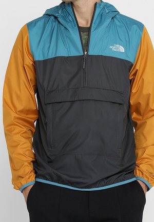 Personne portant un coupe-vent The North Face à blocs de couleurs avec des sections bleu sarcelle, noir et orange, mains dans les poches, avec une fermeture éclair et une poche à l'avant.