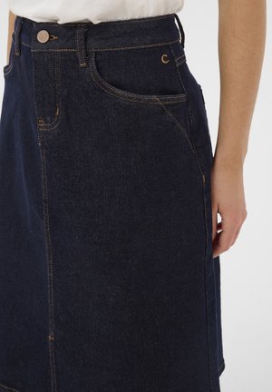 Dunkelblauer Denimrock mit Vordertaschen, Knopfverschluss und sichtbaren orangen Nähten, kombiniert mit einem weißen Oberteil und einer Hand, die seitlich ruht.