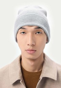 Grå stickad mössa med ribbad textur. Buren över en ljusbrun tröja med kragsdesign. Neutral bakgrund.